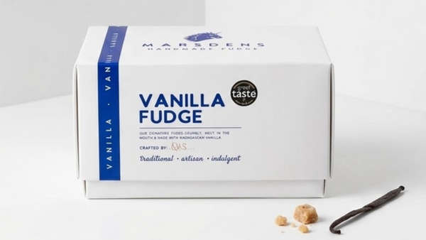 A white box of Marsdens Handcrafted Vanilla Fudge sits on a clean white surface with a few vanilla beans and small fudge pieces placed in front. The box is labeled 'VANILLA FUDGE' with a blue side band and a small circular badge reading 'great taste'.