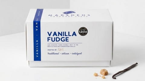 A white box of Marsdens Handcrafted Vanilla Fudge sits on a clean white surface with a few vanilla beans and small fudge pieces placed in front. The box is labeled 'VANILLA FUDGE' with a blue side band and a small circular badge reading 'great taste'.